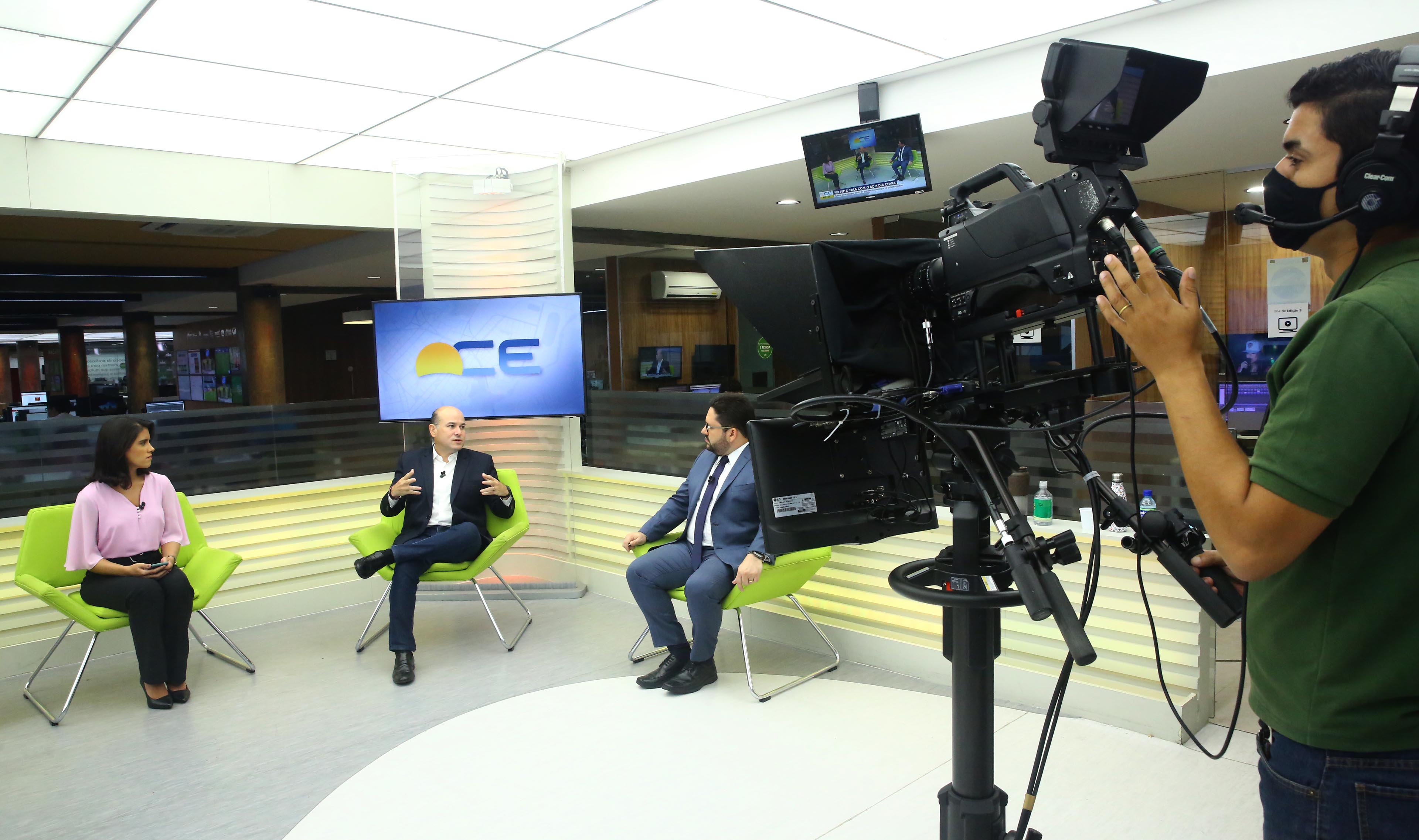 prefeito sentado em estúdio de TV entre os dois apresentadores e cinegrafista ao lado filmando